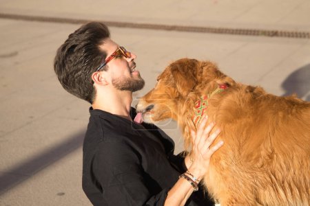 Photo for Young Hispanic man with beard, sunglasses and black shirt, sitting on the floor while his dog licks him in a very affectionate and complicit attitude. Concept animals, dogs, love, pets, golden. - Royalty Free Image