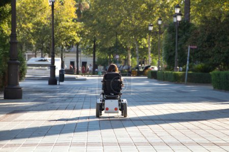 Foto de Mujer con discapacidad, movilidad reducida y baja estatura en silla de ruedas eléctrica caminando por el centro de la ciudad, vista desde atrás. Concepto discapacidad, discapacidad, incapacidad, necesidades especiales. - Imagen libre de derechos