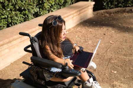 Foto de Mujer discapacitada con movilidad reducida y pequeña estatura en una silla de ruedas eléctrica que trabaja con su computadora portátil al aire libre. Discapacidad conceptual, discapacidad, incapacidad, necesidades especiales, teletrabajo. - Imagen libre de derechos