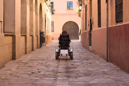 Foto de Mujer discapacitada con movilidad reducida y pequeña estatura en una silla de ruedas eléctrica caminando por una calle solitaria de la ciudad, vista desde atrás. Concepto discapacidad, discapacidad, incapacidad, necesidades especiales. - Imagen libre de derechos