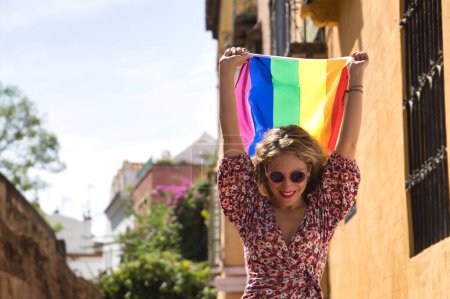 Middle-aged blonde woman, wearing a flowered dress and sunglasses, walking down the street and waving and a gay pride flag. Concept lgtbi, gay, lesbian, pride day.