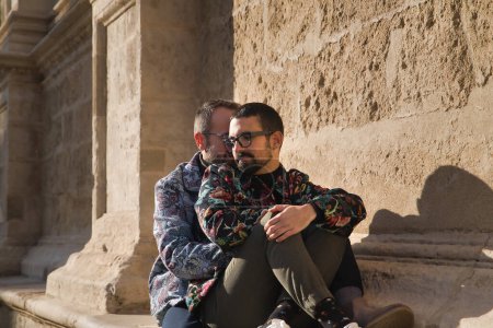Photo for Real marriage of gay couple, sitting on a stone wall while one embraces the other from behind with very affectionate, complicit and happy attitude. Concept lgtb, lgtbiq+, couples, in love, love. - Royalty Free Image