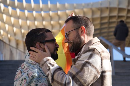 Photo for Real marriage of gay couple, looking at each other while one caresses the other's face and holding a gay pride flag, happy and complicit. Concept lgtb, lgtbiq+, couples, in love, pride, flag. - Royalty Free Image