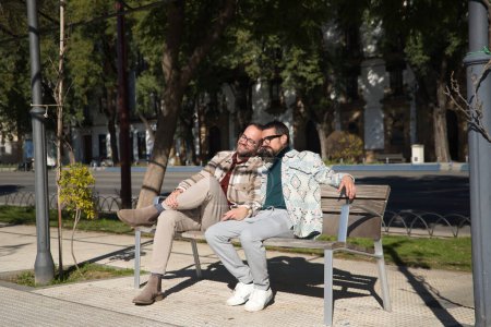 Photo for Real marriage of gay couple, holding hands, heads together and eyes closed, sitting on a wooden bench, complicit and happy. Concept lgtb, lgtbiq+, couples, in love, complicity. - Royalty Free Image