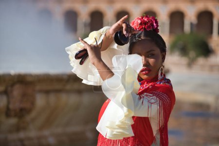 Téléchargez les photos : Jeune femme noire et sud-américaine en costume de flamenco tzigane beige et châle rouge, dansant avec des castagnettes sur une belle place de la ville de Séville en Espagne. Concept dance, folklore, flamenco, art - en image libre de droit