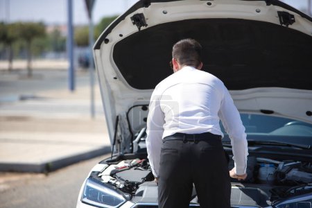 Successful, attractive, young Hispanic man in white shirt, looking at the engine of his white luxury car after it has broken down. Concept, cars, breakdowns, roadside assistance, towing.