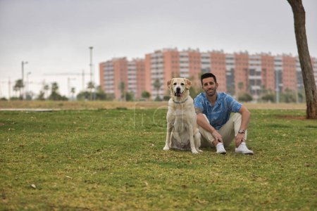 Photo for Young Hispanic man, sitting on the grass next to his dog, both looking at the camera. Concept, dogs, pets, animals, friends. - Royalty Free Image