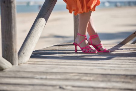 Foto de Detalle de pies de mujer con tacones altos, con tirantes rosas, en una pasarela de madera en la playa. Concepto pies, moda, calzado. - Imagen libre de derechos