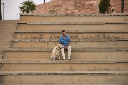 Photo for Young Hispanic man, sitting on big cement stairs next to his dog laughing very happy. Concept, dogs, pets, animals, friends. - Royalty Free Image