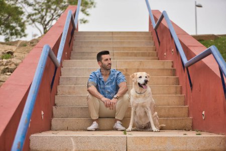 Photo for Young Hispanic male, sitting on stairs next to his dog as they both look in the same direction. Concept, dogs, pets, animals, friends. - Royalty Free Image