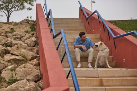 Photo for Young Hispanic man, sitting on stairs next to his dog while talking with him in a loving and tender attitude. Concept, dogs, pets, animals, friends. - Royalty Free Image