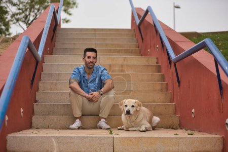 Photo for Young Hispanic male, sitting on stairs next to his dog while looking at the camera. Concept, dogs, pets, animals, friends. - Royalty Free Image