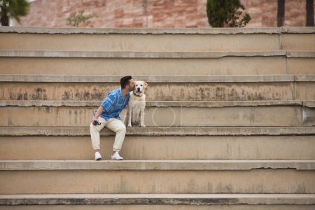 Photo for Young Hispanic man, sitting on big cement stairs giving a kiss to his dog in loving and tender attitude. Concept, dogs, pets, animals, friends. - Royalty Free Image