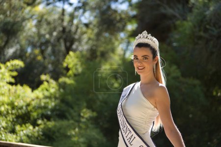 Photo for Portrait of young, beautiful, blonde woman in white suit, diamond crown and beauty pageant winner's sash, posing leaning on a railing. Concept beauty, contests, pageant, fashion. - Royalty Free Image