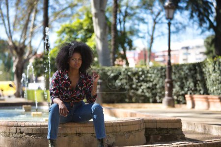 Foto de Mujer joven, guapa y negra de pelo afro, con camisa de flores, jeans y botas, sentada en una fuente jugando con su pelo. Concepto belleza, relajación, peinados. - Imagen libre de derechos