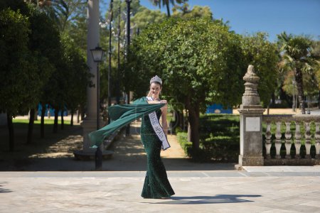 Young, pretty, blonde woman in a green party outfit with sequins, a diamond crown and a beauty pageant winner's sash, twirling so that the air blows her cape. Concept of beauty, fashion, pageant.