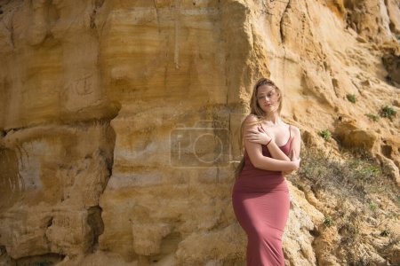 Young, beautiful, blonde woman in a pink dress, sitting on a stone wall, looking at the camera, embracing herself. Concept protection, loving oneself, innocence, tenderness, virginity.