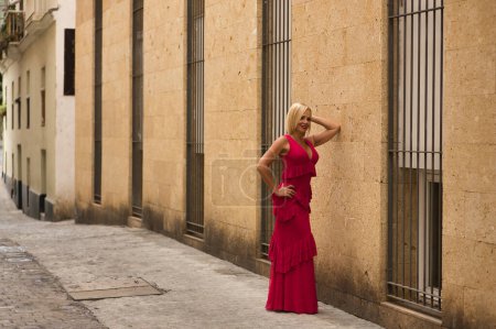 Foto de Mujer madura, guapa, rubia, vestida con un traje de flamenca rosa con volantes y lunares, alegre y sonriente, posando apoyada sobre una pared en una calle solitaria. Concepto belleza, flamenco, madurez. - Imagen libre de derechos