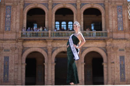 Photo for Young, pretty, blonde woman in a green party dress with sequins, with a diamond crown and beauty pageant winner's sash, posing in the middle of a square. Concept of beauty, fashion, trend, elegance. - Royalty Free Image