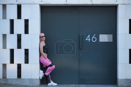 Foto de Atractivo joven gay, fuertemente maquillaje, con pelo rosa, gafas de sol, top de cuero y pantalones, posando apoyado en el marco de una puerta de metal con el número cuarenta y seis. concepto LGTBIQ +, gay, orgullo, maquillaje - Imagen libre de derechos
