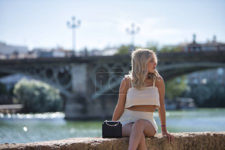 Beautiful young woman, blonde with blue eyes and wearing a white sequined dress, sitting on a stone wall, posing while on vacation in the city, with the Triana bridge in the background.