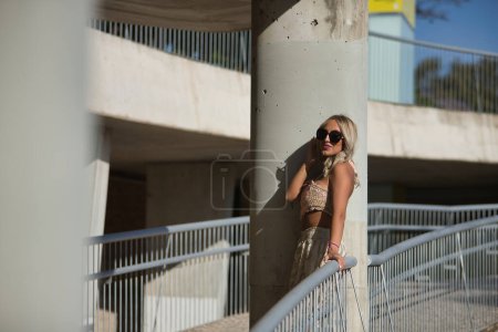 A young, beautiful blonde woman with sunglasses, wearing a crochet top and baggy pants, leaning between a column and a railing posing as a model. Concept of beauty, fashion, trend, model.