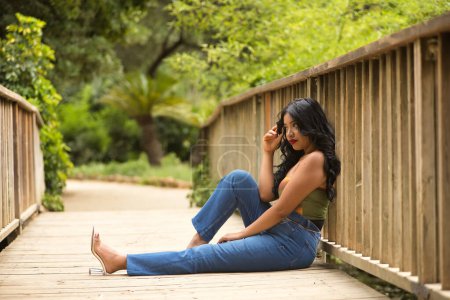 Femme sud-américaine, jeune, belle, brune, avec haut coloré et jeans assis sur un pont en bois, cher et avec un regard perdu. Concept de beauté, mode, tendance, ethnicité, diversité.