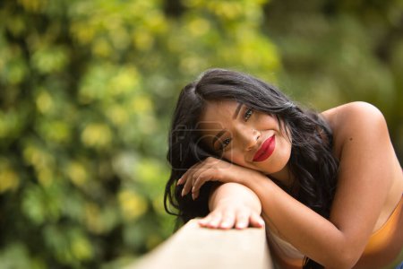 Portrait de femme sud-américaine, jeune, belle, brune, avec un haut coloré et un jean appuyé sur une balustrade en bois au look tendre, pur et rêveur. Concept de beauté, tendance, ethnicité, diversité.