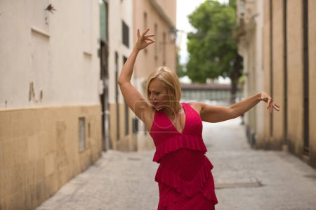 Foto de Mujer madura, guapa, rubia, con traje de flamenca rosa con volantes y lunares, posando como si bailara flamenco. Concepto belleza, moda, tendencia, flamenco, madurez. - Imagen libre de derechos