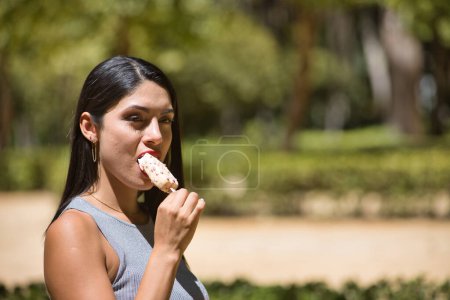 Téléchargez les photos : Portrait de femme sud-américaine, jeune, belle, brune avec haut gris mangeant une crème glacée à la fraise, heureuse et insouciante. Concept beauté, nourriture, été, glace, chaleur. - en image libre de droit