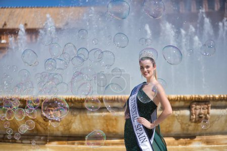 Photo for Young, pretty, blonde woman in a green party outfit with sequins, with a diamond crown and beauty pageant winner's sash, posing next to a fountain surrounded by soap bubbles. - Royalty Free Image