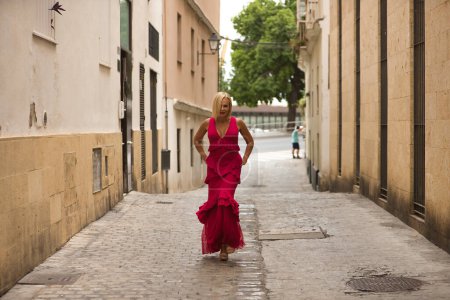 Foto de Mujer madura, guapa, rubia, con traje de flamenca rosa con volantes y lunares, alegre y sonriente, caminando y vistiendo el vestido. Concepto belleza, moda, tendencia, flamenco, madurez. - Imagen libre de derechos