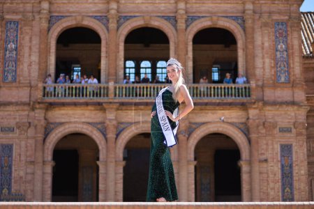 Photo for Young, pretty, blonde woman in a green party dress with sequins, with a diamond crown and beauty pageant winner's sash, posing in the middle of a square. Concept of beauty, fashion, trend, elegance. - Royalty Free Image