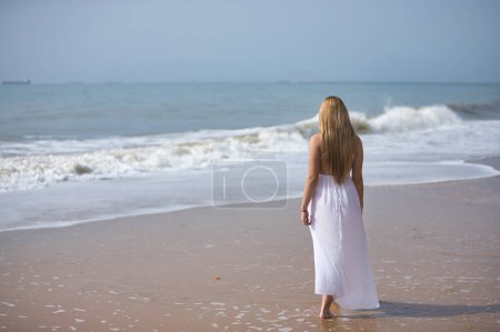 Jeune, belle femme blonde en robe blanche, marchant sur une plage solitaire, détendue et calme, vue du dos. Concept paix, tranquillité, solitude, se retrouver.