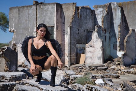 Mujer sudamericana, joven, guapa, morena con lencería y alas negras, posando agachada en medio de las ruinas de un edificio. Concepto ángeles, belleza, disfraces, alas, halloween.