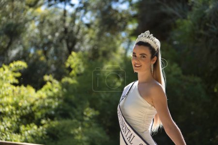 Portrait of young, beautiful, blonde woman in white suit, diamond crown and beauty pageant winner's sash, posing leaning on a railing. Concept beauty, contests, pageant, fashion.