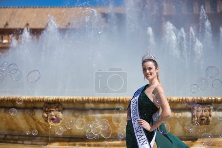 Photo for Young, pretty, blonde woman in a green party outfit with sequins, with a diamond crown and beauty pageant winner's sash, posing next to a fountain surrounded by soap bubbles. - Royalty Free Image