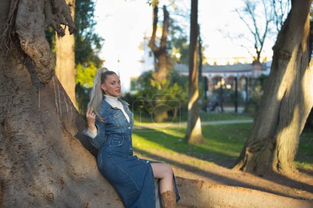 Frau, jung, hübsch, blond, mit blauen Augen und Jeanskleid, berührt ihre Haare kokett und blickt in die Kamera, die auf dem Stamm eines großen Baumes sitzt und die Sonnenstrahlen beim Sonnenuntergang empfängt..
