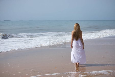 Jeune, belle femme blonde en robe blanche, marchant sur une plage solitaire, détendue et calme, vue du dos. Concept paix, tranquillité, solitude, se retrouver.