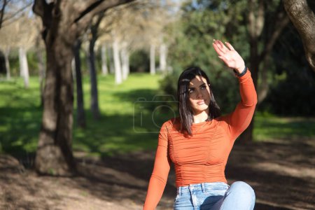 Eine junge, schöne Frau in orangefarbenem T-Shirt und Jeans, die ihr Gesicht mit der Handfläche vor den Sonnenstrahlen verdeckt, entspannt und ruhig auf einem riesigen Stein in einem Park sitzend. Konzept Sonne, Schutz.