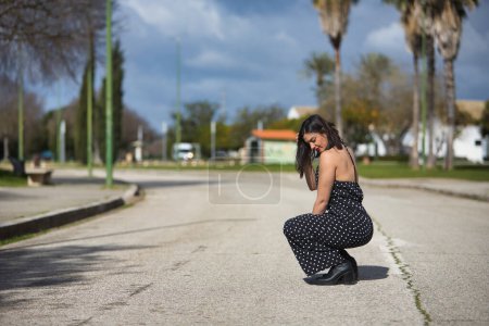 Photo for A young, beautiful woman in a black dress with white polka dots, crouched down, sad and alone, in the middle of a road. Concept loneliness, sorrow, beauty, fashion. - Royalty Free Image
