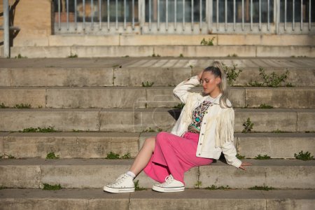 Young, pretty, blonde, with her eyes closed, white fringed jacket and pink skirt, relaxed and calm sitting on some steps. Concept of beauty, fashion, millennial, trend.
