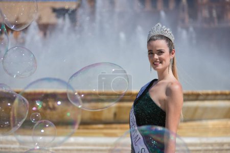 Photo for Portrait of young, pretty, blonde woman in green sequined party outfit, with diamond crown and beauty pageant winner's sash, posing next to a fountain surrounded by soap bubbles. - Royalty Free Image