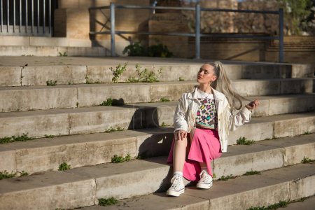 Young, pretty, blonde woman, with her eyes closed, white fringed jacket and pink skirt, touching her hair, relaxed and calm sitting on some steps. Concept of beauty, fashion, millennial, trend.