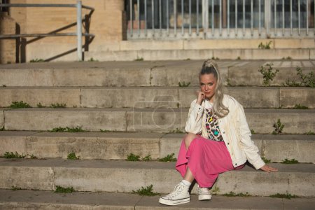 Young, pretty, blonde woman, with blue eyes, white fringed jacket and pink skirt, with her hand on her chin looking at the camera, relaxed and calm sitting on some steps.