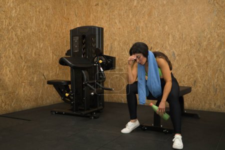 Young, beautiful, brunette woman with yellow top, black leggings and towel around her neck, tired, exhausted, depressed and frustrated, sitting on a bench in the gym. Fitness concept, gymnastics.