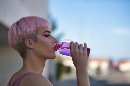 Portrait of young, attractive, gay, heavily makeup man with pink hair and top, drinking water from a bottle on a hot afternoon. LGTBIQ+ concept, gay, pride, makeup, fashion, trend.
