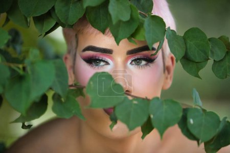 Porträt eines jungen attraktiven homosexuellen Mannes, mit grünen Augen, rosa Haaren, stark geschminkt, der durch die Blätter und Äste einiger grüner Rebstöcke blickt. LGTBIQ + Konzept, Homosexuell, Stolz, Make-up, Mode.