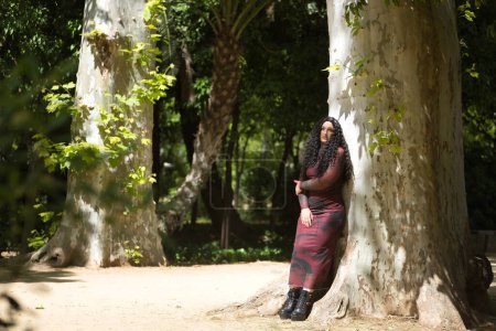 Young, pretty, alternative and chubby woman, wearing a red dress, loving herself and connecting with nature leaning on the trunk of a large tree. Millennial, plump, independent, different.