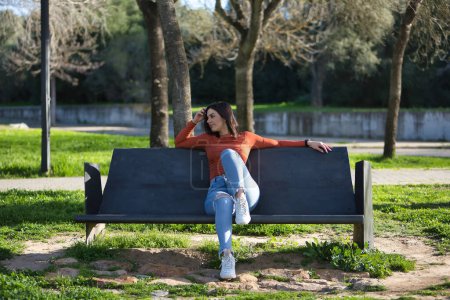 Eine junge, schöne Frau in orangefarbenem T-Shirt und Jeans, abgelenkt und in ihren Gedanken versunken, sitzt auf einer Metallbank in einem Park. Modekonzept, Schönheit, Ruhe, Entspannung.